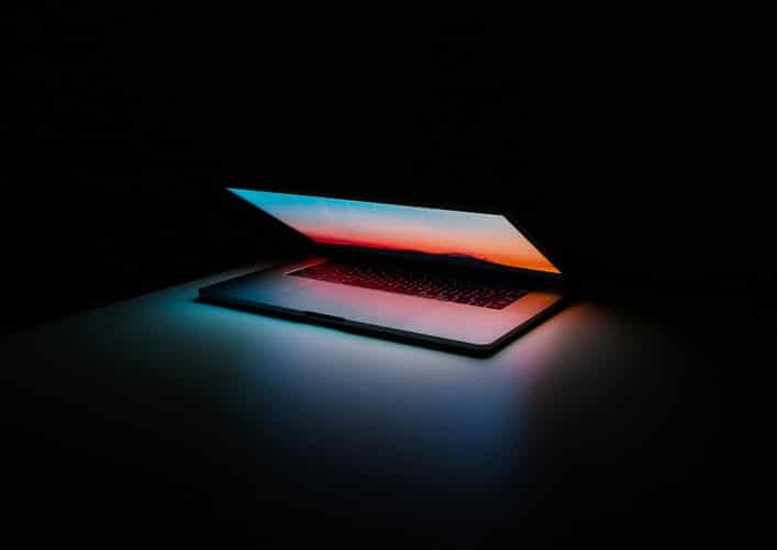 notebook semiaberto refletindo luzes azuis, rosa e laranja em fundo preto