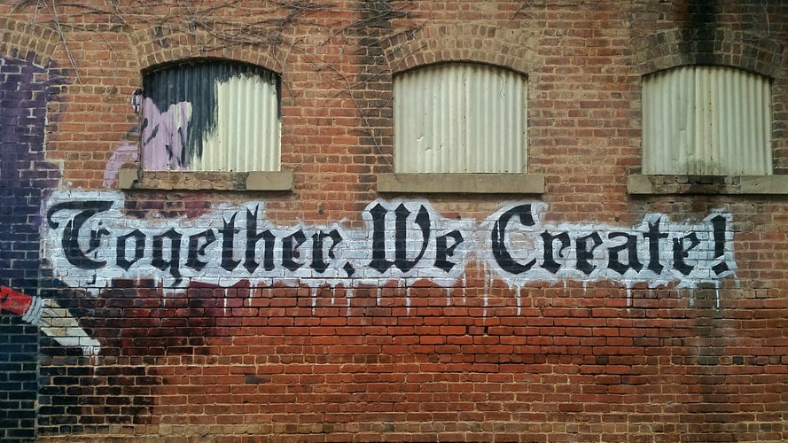 Parede de tijolos onde está escrito em letras pretas com contorno branco a frase "together, we create!"